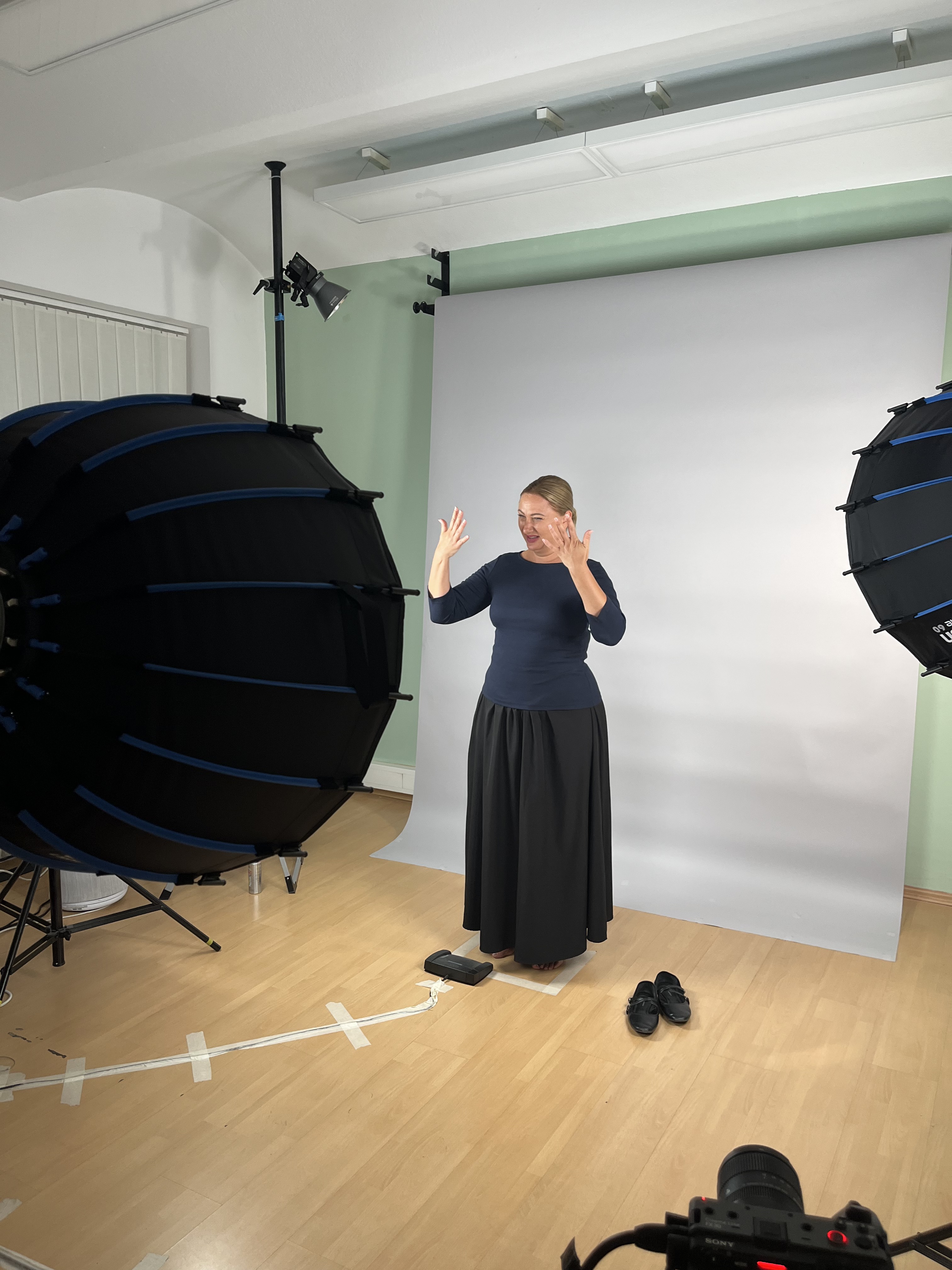 Eine Darstellerin gebärdet mit beiden Händen vor einem hellen Studiohintergrund. Im Vordergrund stehen Kamera und Studiolichter.