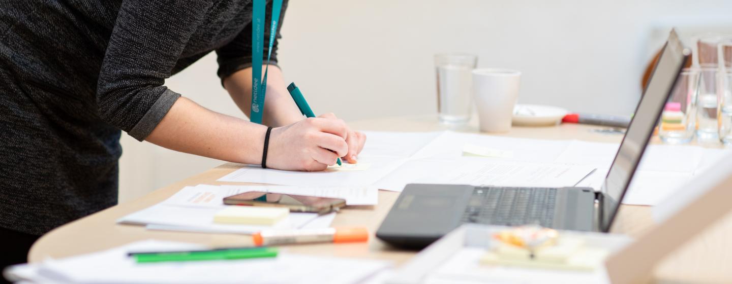 Person writing with a pen on paper next to a laptop