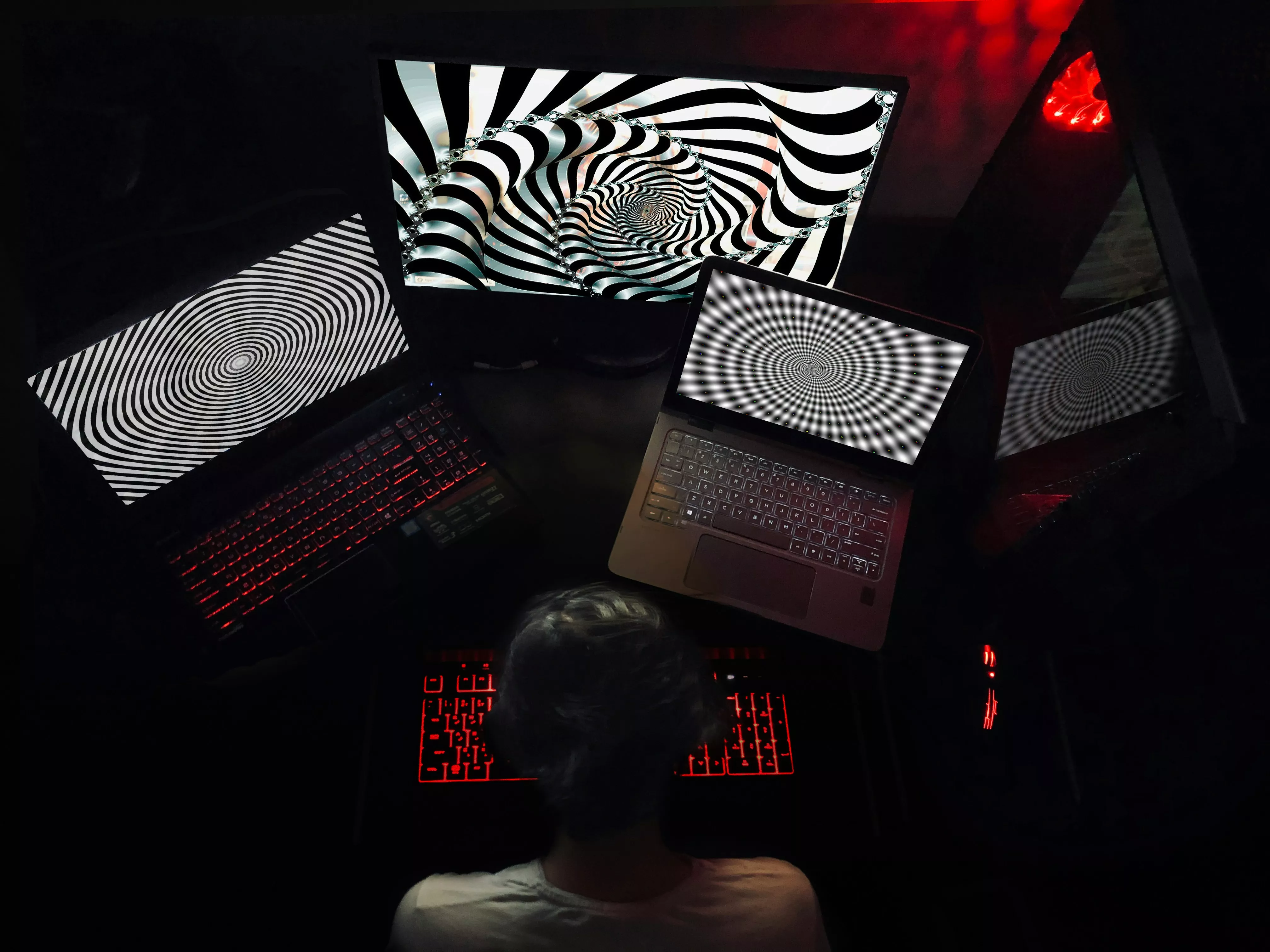 Eine Person sitzt in einem dunklen Raum vor mehreren Laptops und Monitoren, die hypnotische schwarz-weiße Spiralmuster anzeigen, mit rot beleuchteter Tastatur.