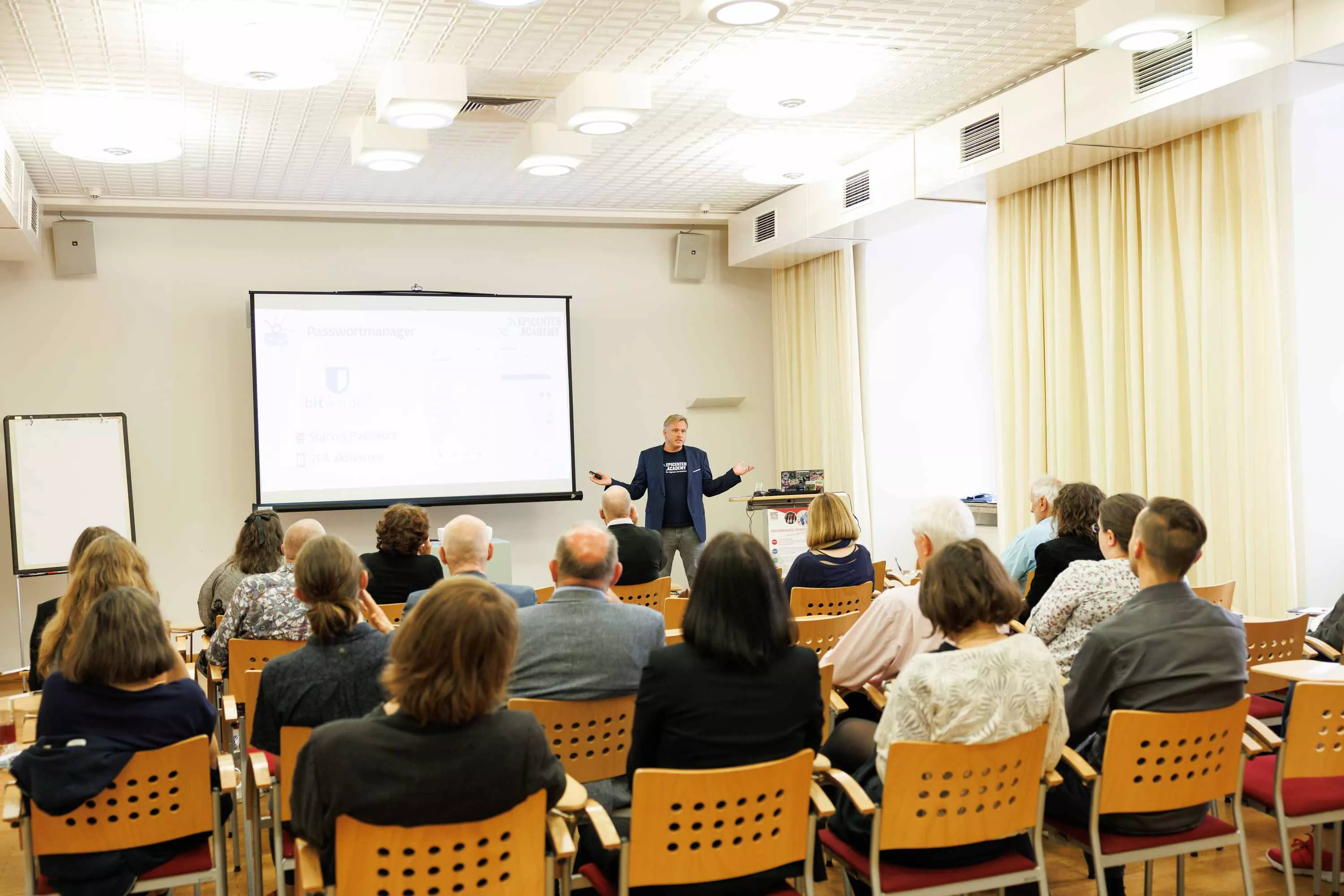 Daniel Lohninger steht vor einem Publikum am OSGS Forum in einem hellen Seminarraum und hält einen Vortrag zum Thema Datenschutz und IT-Sicherheit. Auf der Leinwand hinter ihm ist das Logo von „Bitwarden“ sowie der Schriftzug „epicenter.academy“ zu sehen. Die Zuhörer:innen verfolgen aufmerksam die Präsentation. Der Raum ist mit Holzstühlen, Deckenleuchten und großen Fenstern mit Vorhängen ausgestattet. Ein Flipchart steht ungenutzt am Rand. Copyright:Sima Prodinger