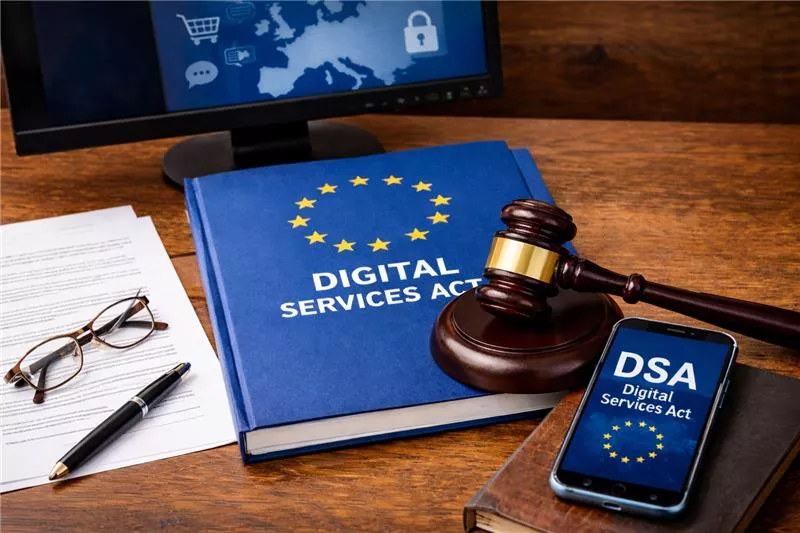 Auf einem Holztisch liegt ein blaues Buch mit der Aufschrift „DIGITAL SERVICES ACT“ und einem Kreis gelber Sterne. Darauf liegt ein Richterhammer. Rechts daneben befindet sich ein Smartphone mit der Anzeige „DSA – Digital Services Act“. Links liegen Dokumente mit einer Brille und einem Stift. Im Hintergrund ist ein Monitor mit einer Europa-Karte und digitalen Symbolen zu sehen.