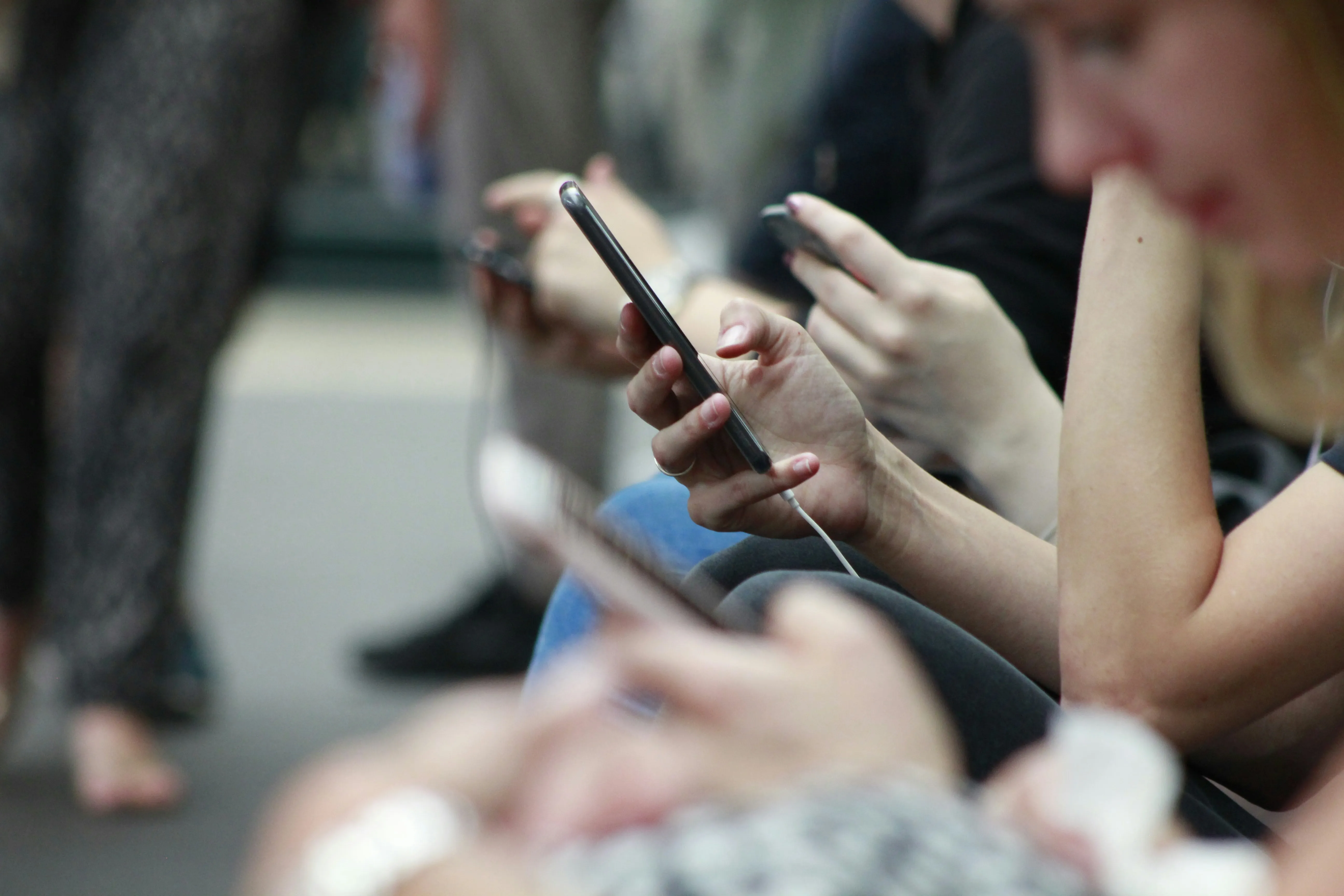 Mehrere junge Menschen sitzen nebeneinander und nutzen Smartphones; der Fokus liegt auf einer Hand mit Handy im Vordergrund, die Gesichter sind unscharf. Das Bild symbolisiert die alltägliche Nutzung sozialer Medien und den Bedarf an altersgerechten Schutz- und Privatsphäre-Einstellungen.