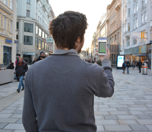 Person von hinten, die vor einer Fußgängerzone steht und ein Smartphone in die Höhe hält.