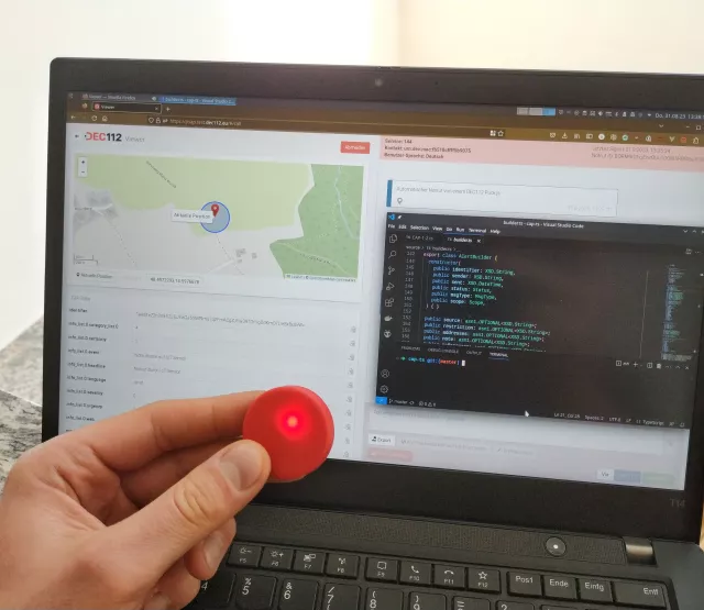 Laptop mit laufendem DEC112 Viewer. Im Vordergrund eine Hand mit einem Puck.js, welcher rot leuchtet.