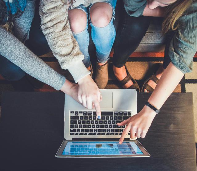 Man sieht einen Laptop von oben und die Knie und Hände von drei Personen, die auf diesen Laptop zeigen 