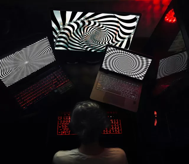 Eine Person sitzt in einem dunklen Raum vor mehreren Laptops und Monitoren, die hypnotische schwarz-weiße Spiralmuster anzeigen, mit rot beleuchteter Tastatur.