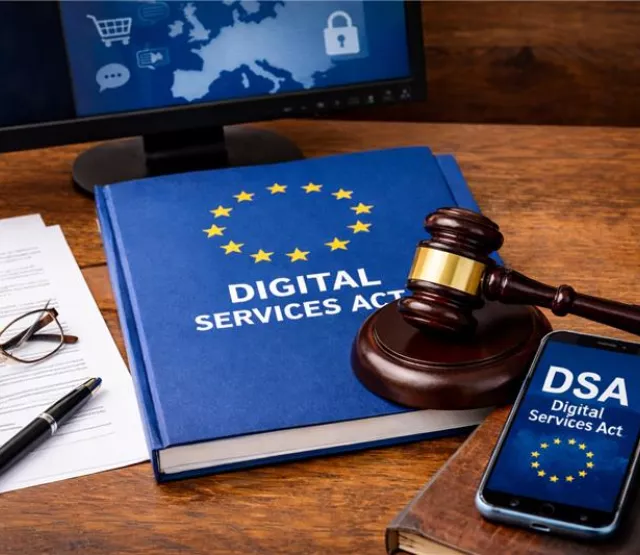 Auf einem Holztisch liegt ein blaues Buch mit der Aufschrift „DIGITAL SERVICES ACT“ und einem Kreis gelber Sterne. Darauf liegt ein Richterhammer. Rechts daneben befindet sich ein Smartphone mit der Anzeige „DSA – Digital Services Act“. Links liegen Dokumente mit einer Brille und einem Stift. Im Hintergrund ist ein Monitor mit einer Europa-Karte und digitalen Symbolen zu sehen.