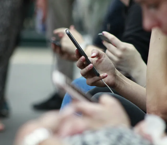 Mehrere junge Menschen sitzen nebeneinander und nutzen Smartphones; der Fokus liegt auf einer Hand mit Handy im Vordergrund, die Gesichter sind unscharf. Das Bild symbolisiert die alltägliche Nutzung sozialer Medien und den Bedarf an altersgerechten Schutz- und Privatsphäre-Einstellungen.