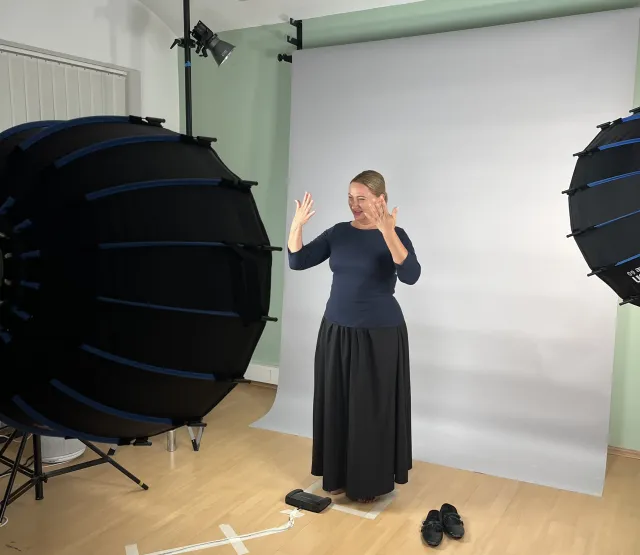 Eine Darstellerin gebärdet mit beiden Händen vor einem hellen Studiohintergrund. Im Vordergrund stehen Kamera und Studiolichter.