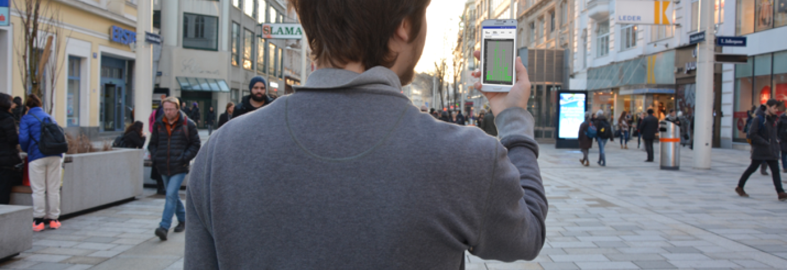 Person von hinten, die vor einer Fußgängerzone steht und ein Smartphone in die Höhe hält.