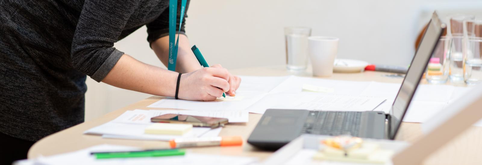 Person writing with a pen on paper next to a laptop