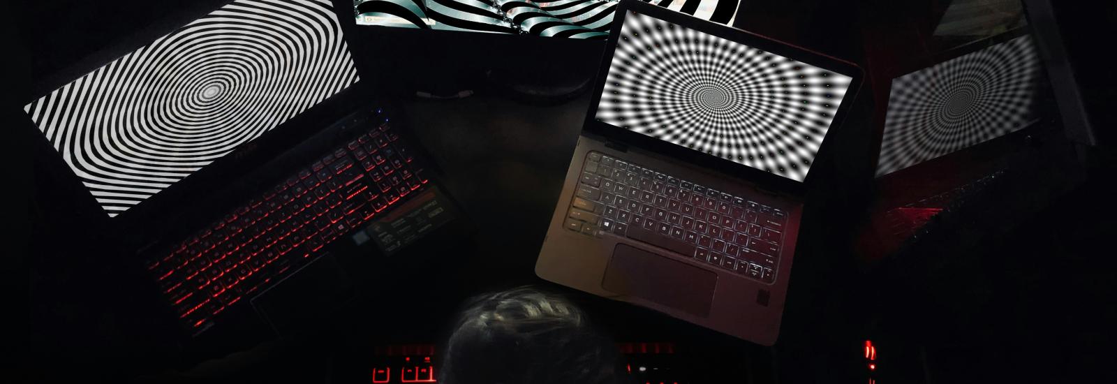 Eine Person sitzt in einem dunklen Raum vor mehreren Laptops und Monitoren, die hypnotische schwarz-weiße Spiralmuster anzeigen, mit rot beleuchteter Tastatur.