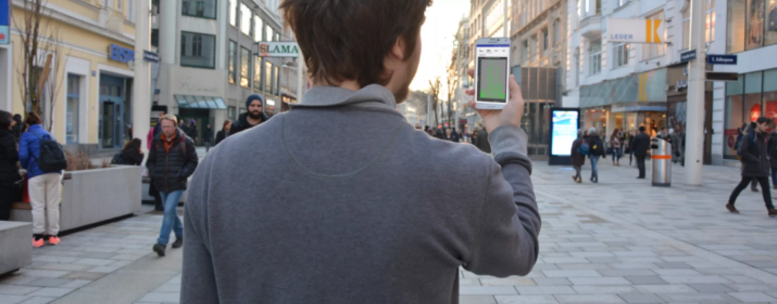 Person von hinten, die vor einer Fußgängerzone steht und ein Smartphone in die Höhe hält.