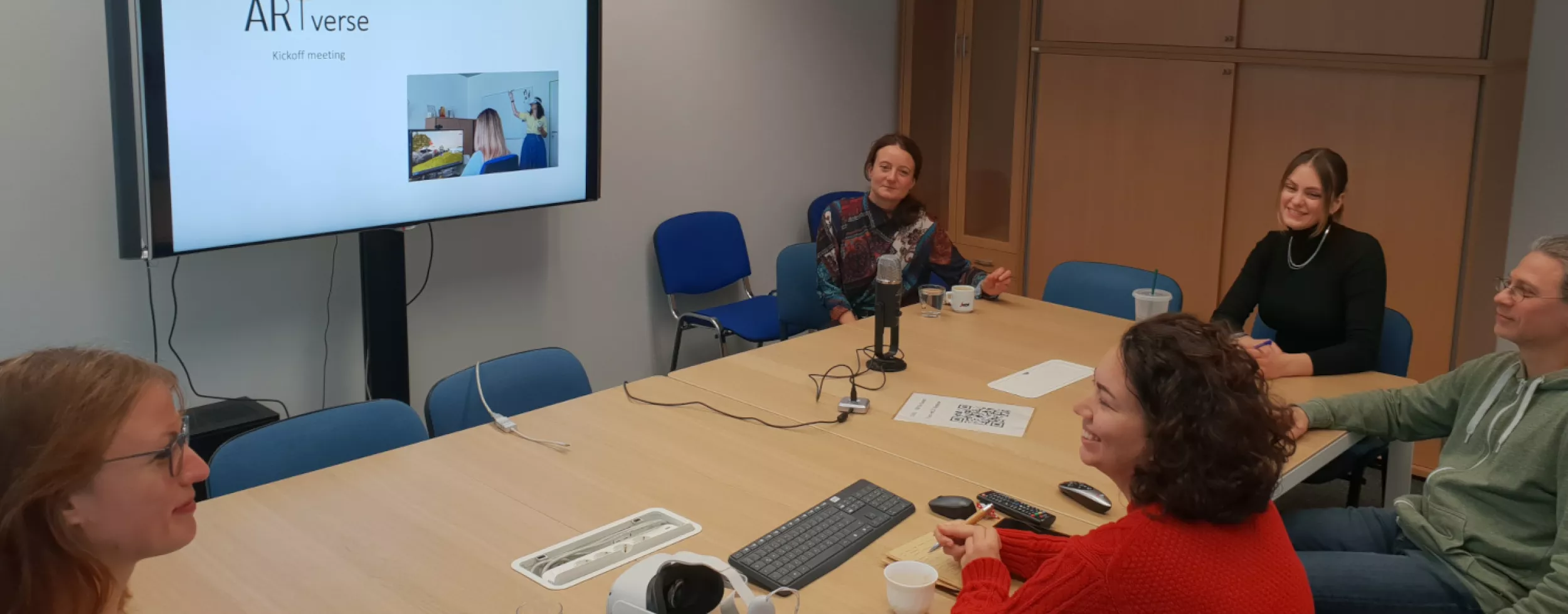 ARTverse's five team members sit around a meeting table while watching a presentation on a television screen.