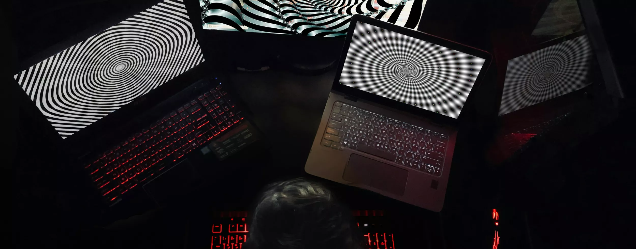 Eine Person sitzt in einem dunklen Raum vor mehreren Laptops und Monitoren, die hypnotische schwarz-weiße Spiralmuster anzeigen, mit rot beleuchteter Tastatur.