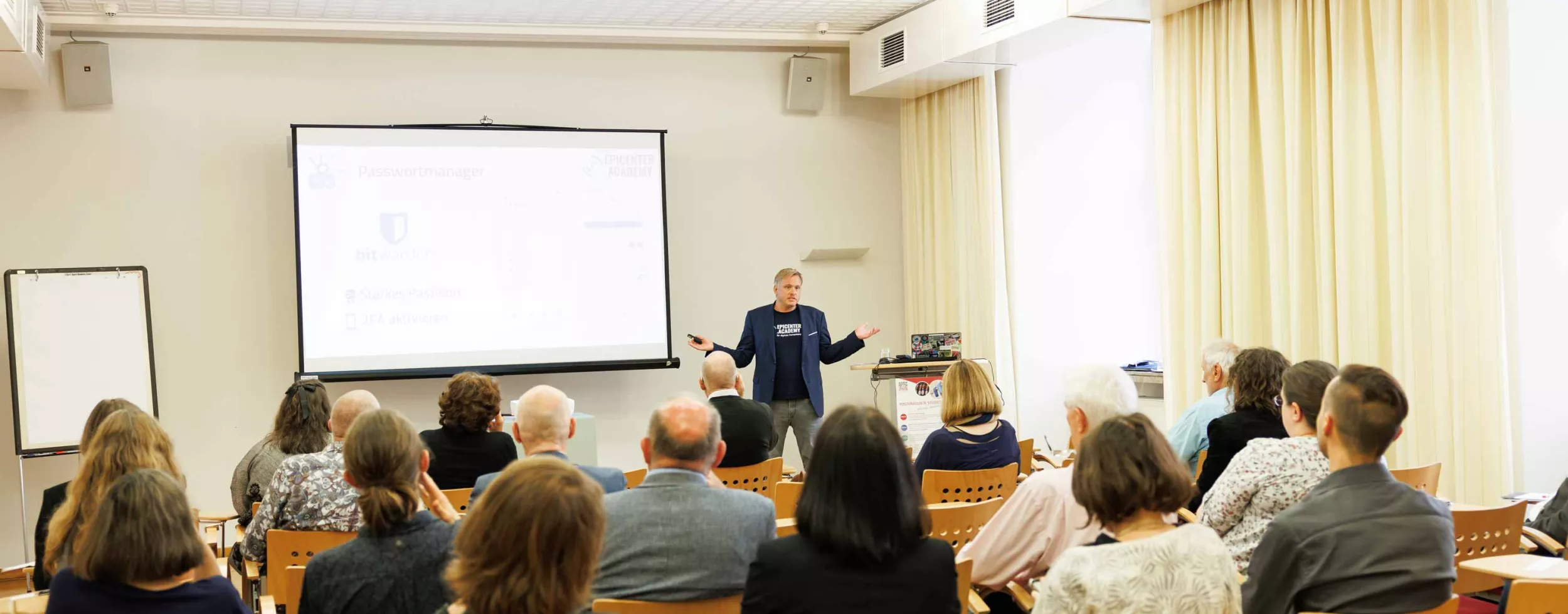Daniel Lohninger steht vor einem Publikum am OSGS Forum in einem hellen Seminarraum und hält einen Vortrag zum Thema Datenschutz und IT-Sicherheit. Auf der Leinwand hinter ihm ist das Logo von „Bitwarden“ sowie der Schriftzug „epicenter.academy“ zu sehen. Die Zuhörer:innen verfolgen aufmerksam die Präsentation. Der Raum ist mit Holzstühlen, Deckenleuchten und großen Fenstern mit Vorhängen ausgestattet. Ein Flipchart steht ungenutzt am Rand. Copyright:Sima Prodinger