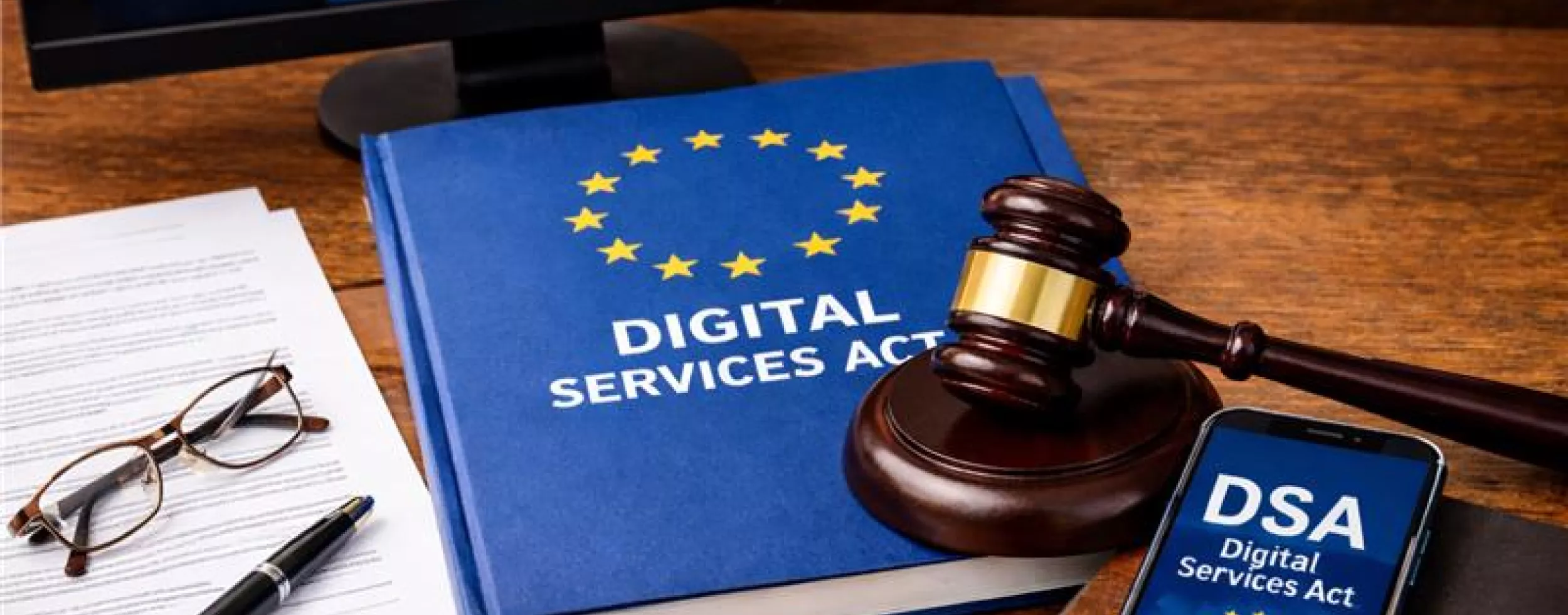 Auf einem Holztisch liegt ein blaues Buch mit der Aufschrift „DIGITAL SERVICES ACT“ und einem Kreis gelber Sterne. Darauf liegt ein Richterhammer. Rechts daneben befindet sich ein Smartphone mit der Anzeige „DSA – Digital Services Act“. Links liegen Dokumente mit einer Brille und einem Stift. Im Hintergrund ist ein Monitor mit einer Europa-Karte und digitalen Symbolen zu sehen.