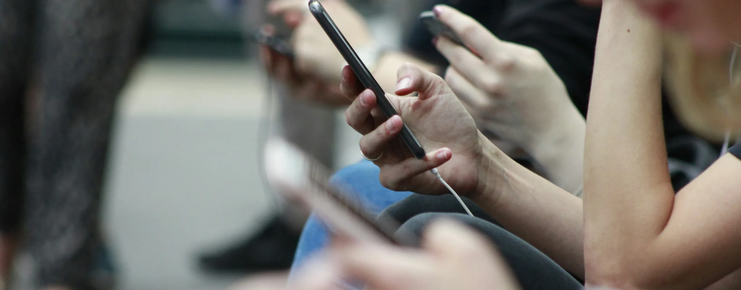 Mehrere junge Menschen sitzen nebeneinander und nutzen Smartphones; der Fokus liegt auf einer Hand mit Handy im Vordergrund, die Gesichter sind unscharf. Das Bild symbolisiert die alltägliche Nutzung sozialer Medien und den Bedarf an altersgerechten Schutz- und Privatsphäre-Einstellungen.