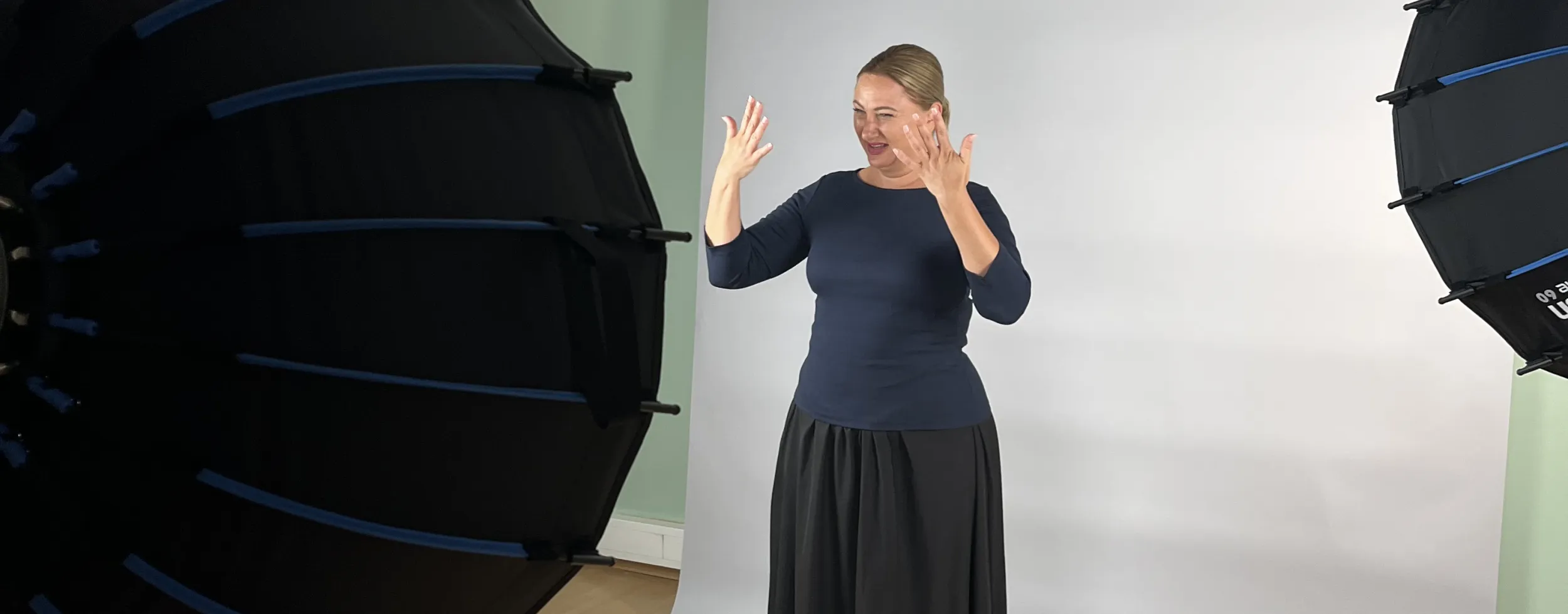 Eine Darstellerin gebärdet mit beiden Händen vor einem hellen Studiohintergrund. Im Vordergrund stehen Kamera und Studiolichter.
